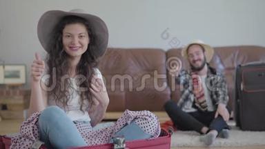肖像快乐的男人和女人坐在地板上，然后在家里旅行。 坐在手提箱里的可爱女人和一个